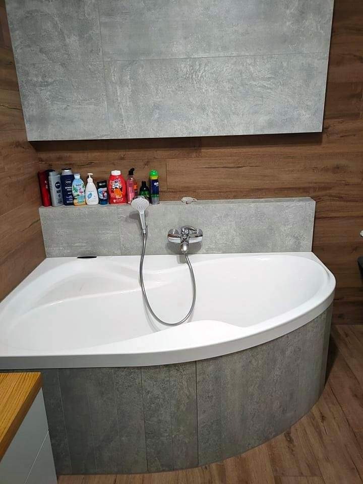 Narożna wanna obudowana szarymi płytkami imitującymi kamień, z baterią i słuchawką prysznicową, na tle drewnopodobnych paneli ściennych i półki z kosmetykami.