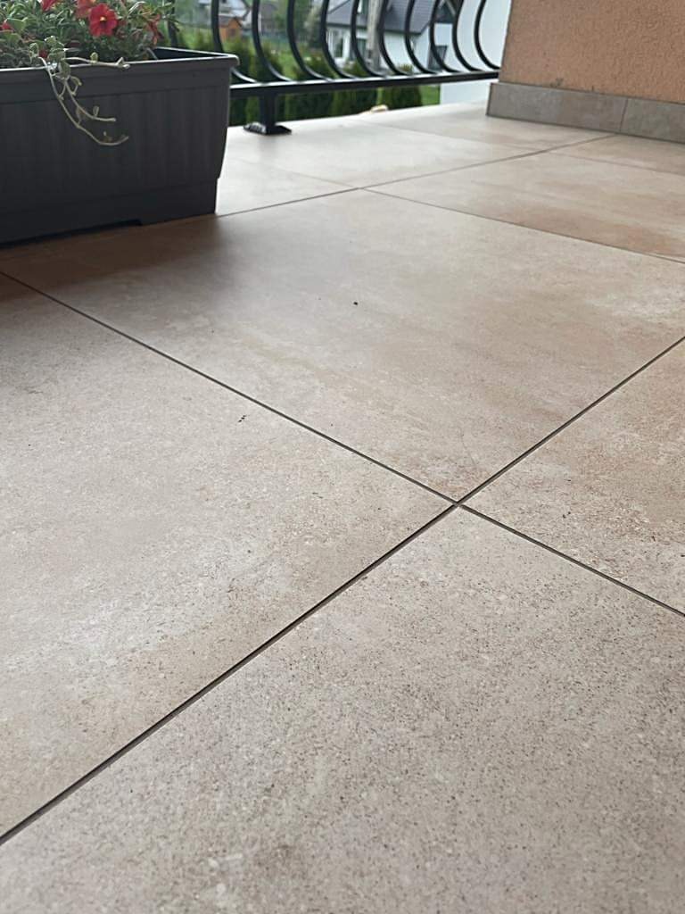 Beżowe płytki gresowe ułożone na balkonie, widoczna donica z kwiatami i metalowa balustrada w tle.