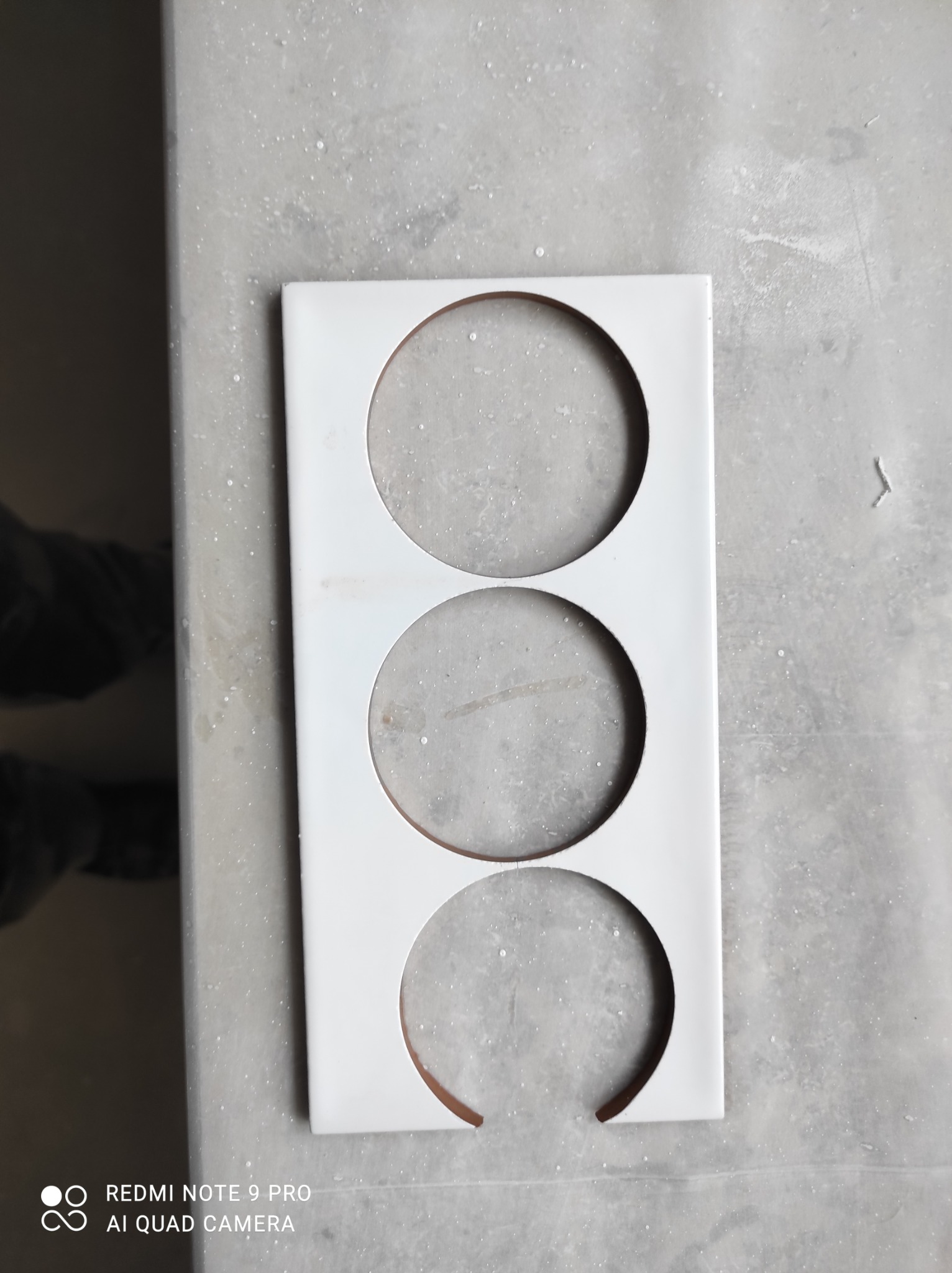 Biała płytka ceramiczna z trzema okrągłymi otworami, leżąca na szarej, lekko zabrudzonej powierzchni, prawdopodobnie przygotowana do montażu w łazience lub kuchni.