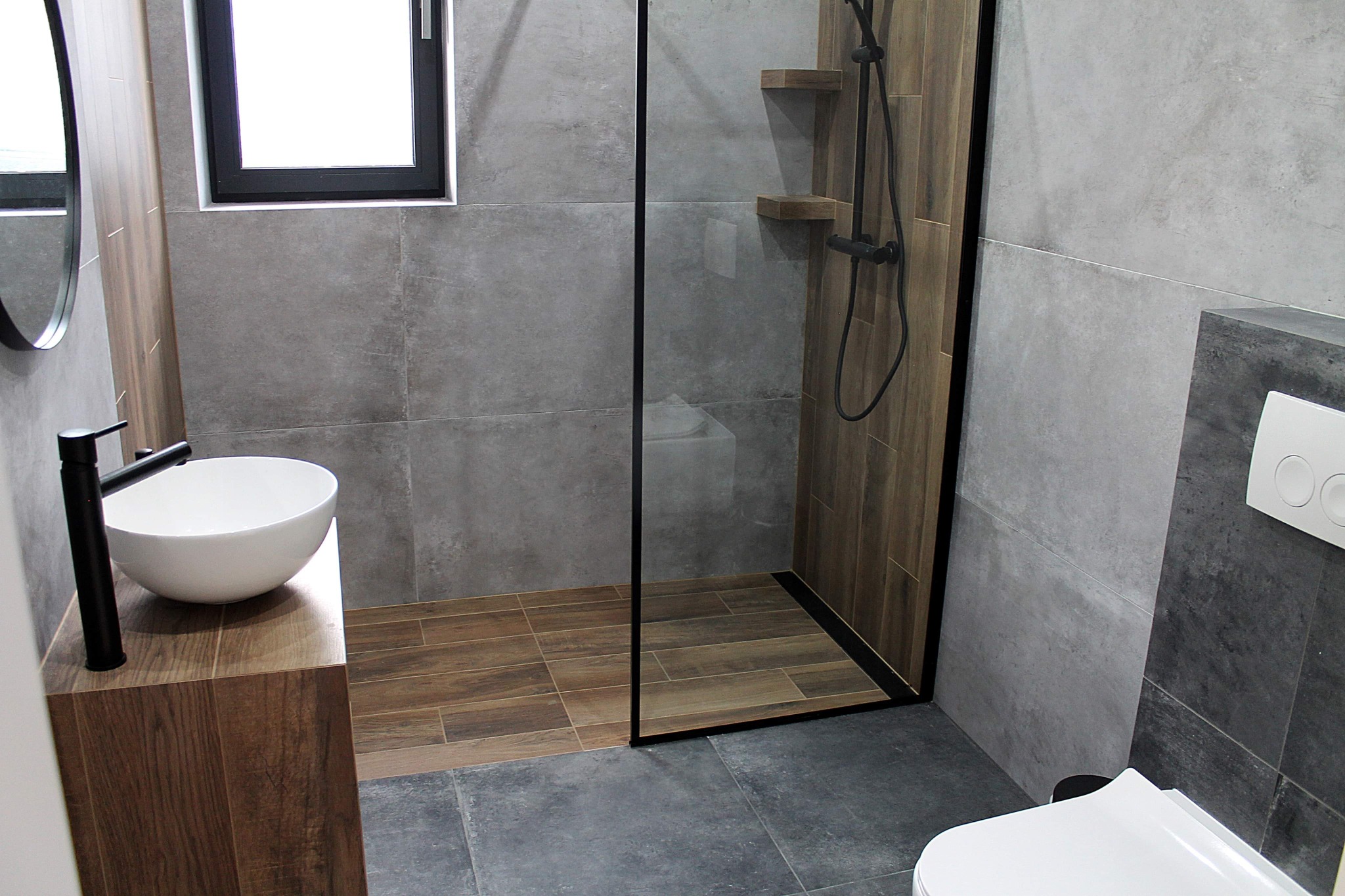 Nowoczesna łazienka z szarymi płytkami imitującymi beton, drewnopodobnymi panelami w strefie prysznica walk-in, umywalką nablatową na drewnianej szafce i czarną armaturą.