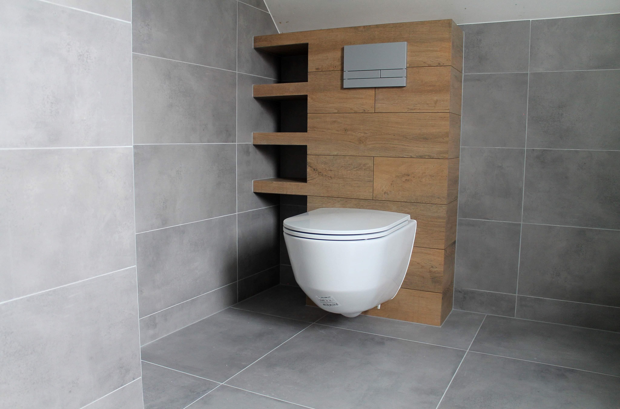 Wisząca toaleta z drewnianą zabudową i półkami we wnęce, szare płytki podłogowe i ścienne, minimalistyczny styl, przycisk spłukujący w kolorze srebrnym.