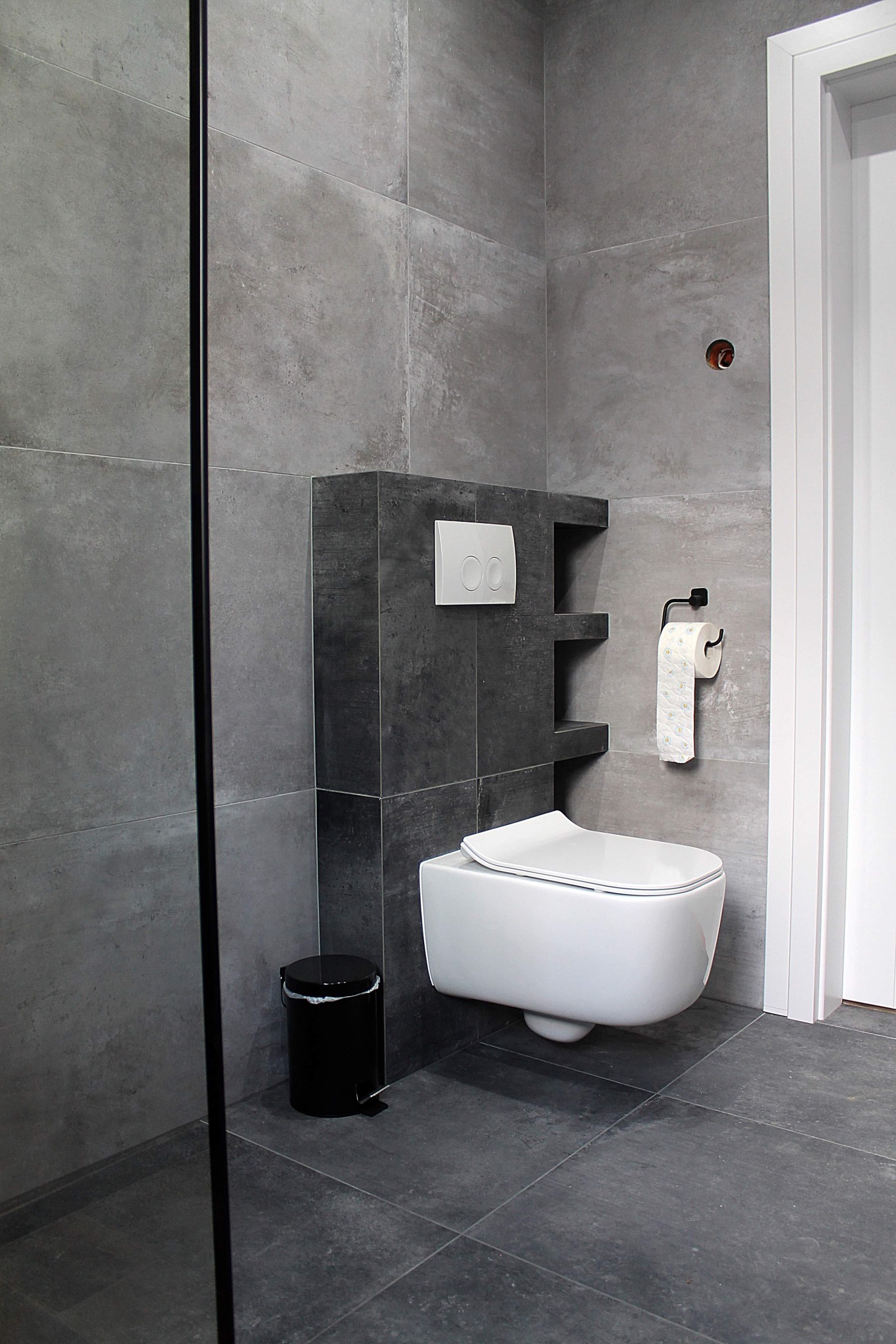 Nowoczesna łazienka z szarymi płytkami imitującymi beton, wisząca miska WC, czarny kosz na śmieci i uchwyt na papier toaletowy.