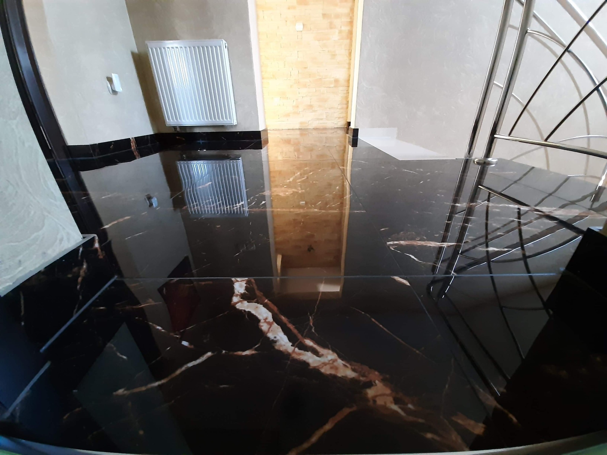 Błyszcząca, czarna podłoga z żyłkowaniem w odcieniach brązu i bieli, odbijająca elementy wnętrza: grzejnik, drzwi i fragment schodów z metalową balustradą.