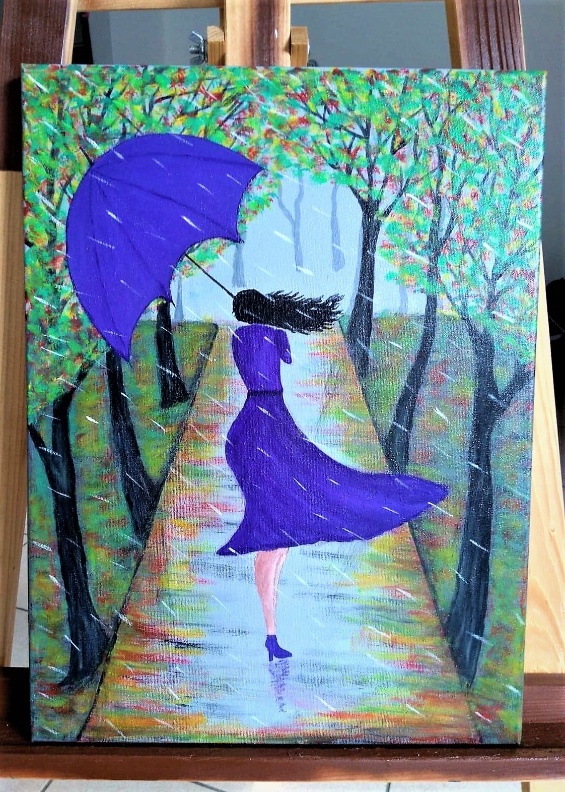 Obraz przedstawia kobietę w fioletowej sukience i z parasolem, idącą ścieżką w deszczowy dzień. Drzewa po bokach ścieżki mają zielone i pomarańczowe liście. Obraz stoi na drewnianej sztaludze.