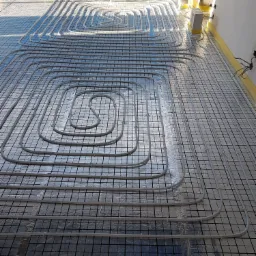 Instalacja ogrzewania podłogowego z rur rozprowadzonych spiralnie na folii z siatką, widoczne żółte elementy izolacyjne przy ścianach i betonowe bloczki stabilizujące.