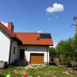 Dom jednorodzinny z czerwoną dachówką, panelami słonecznymi na dachu i drewnianą bramą garażową, widok z ogrodu w słoneczny dzień.