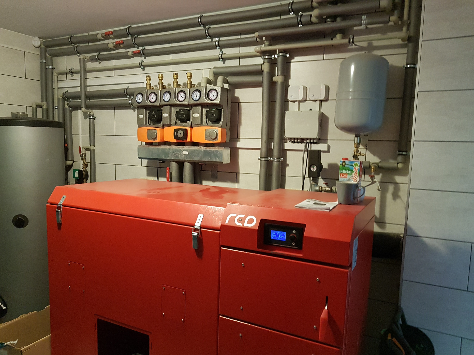 Czerwony piec na pellet marki RED w kotłowni, widoczne liczne rury instalacyjne, manometry, zbiornik wyrównawczy oraz karton mleka i kubek na piecu.