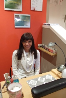 Uśmiechnięta kobieta siedzi za biurkiem w salonie, na którym znajdują się akcesoria do stylizacji paznokci, a na ścianie wiszą certyfikaty.