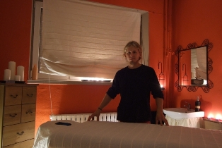 Wnętrze pomarańczowego gabinetu z łóżkiem do masażu, blond kobieta w ciemnej bluzce stoi obok łóżka, w tle okno z roletą i komoda z zapalonymi świecami, na ścianie lustro w ozdobnej ramie.