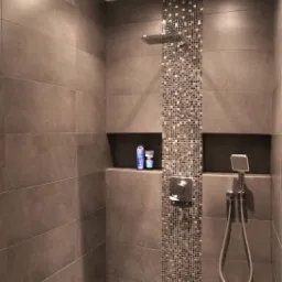 Nowoczesna kabina prysznicowa z szarymi płytkami wielkoformatowymi i mozaiką w pionowym pasie, wnęki na kosmetyki, deszczownica i słuchawka prysznicowa.