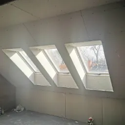 Poddasze z trzema oknami dachowymi, obłożone płytami gipsowo-kartonowymi, widoczne ślady po szpachlowaniu na ścianie z lewej strony, na podłodze butelki i torba.