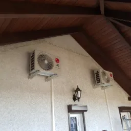 Dwa klimatyzatory LG zamontowane na zewnątrz budynku z drewnianym zadaszeniem, widoczna lampa ścienna i drabina oparta o okno z roletą.