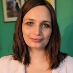 Portret kobiety z brązowymi włosami i zielonymi oczami, ubrana w białą marynarkę, na tle zielonej ściany z obrazami.