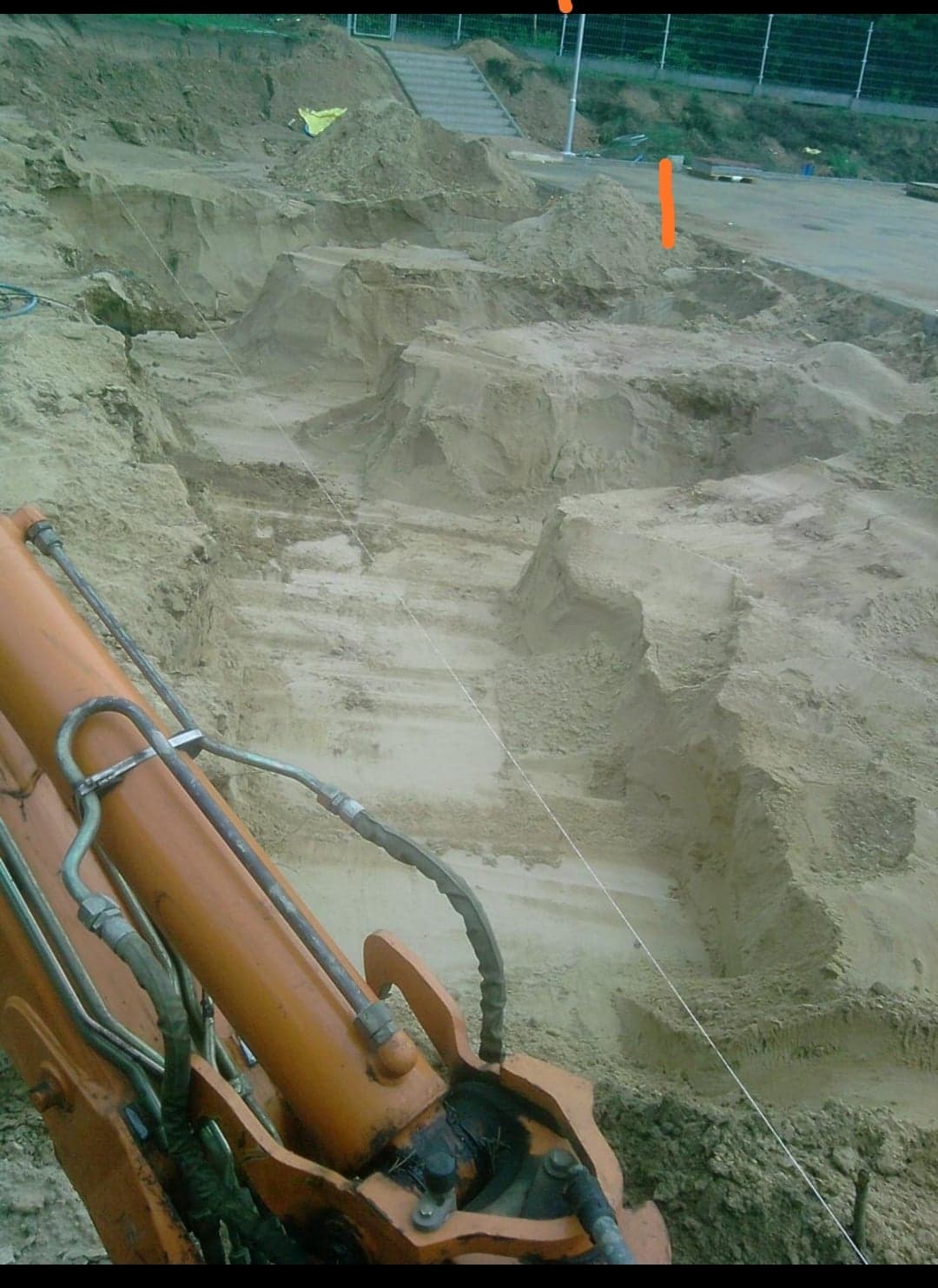 Pomarańczowy wysięgnik koparki na pierwszym planie, w tle wykopany teren z warstwami ziemi, widoczna linka wyznaczająca linię, schody w tle.