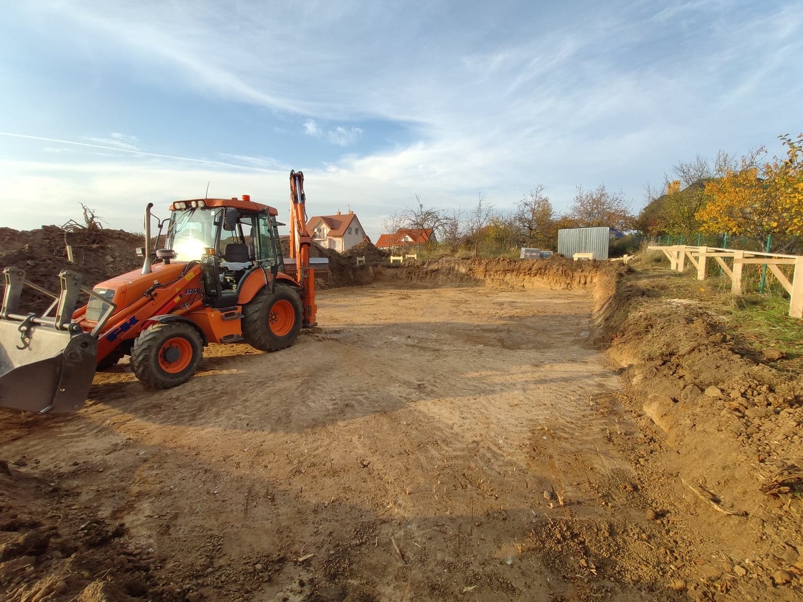 Pomarańczowa koparko-ładowarka stoi obok wykopu pod fundamenty, z widocznymi śladami opon na piaszczystej glebie i drewnianymi podporami w tle, pod błękitnym niebem z delikatnymi chmurami.