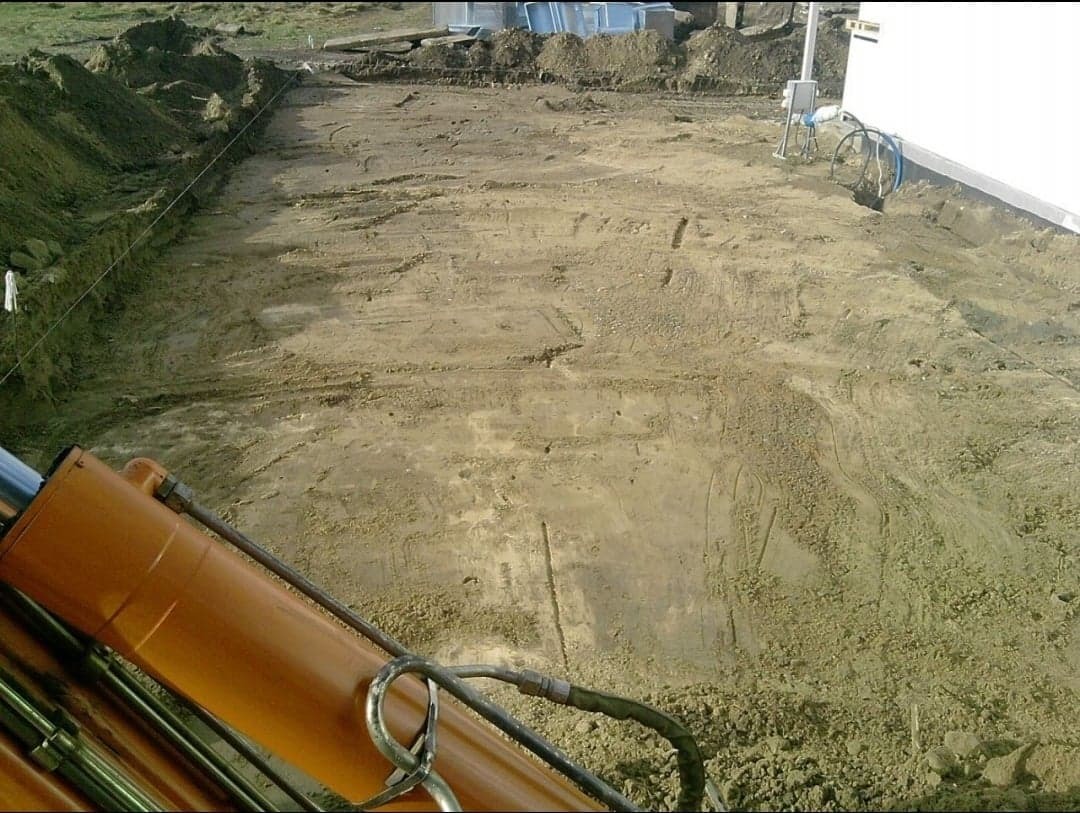 Przygotowany teren pod budowę z widocznymi śladami opon i elementami pomarańczowego ramienia koparki w dolnej części kadru, obok biała ściana budynku.