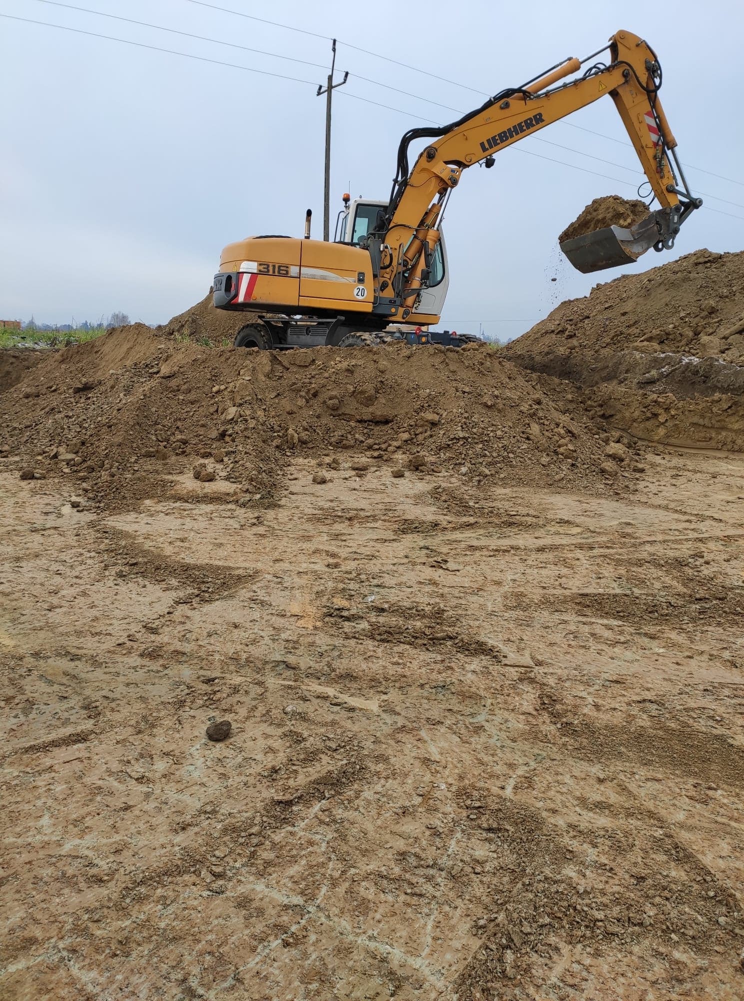Żółta koparka Liebherr 316 nabiera ziemię z pryzmy na tle pochmurnego nieba, widoczne ślady opon na ziemi.
