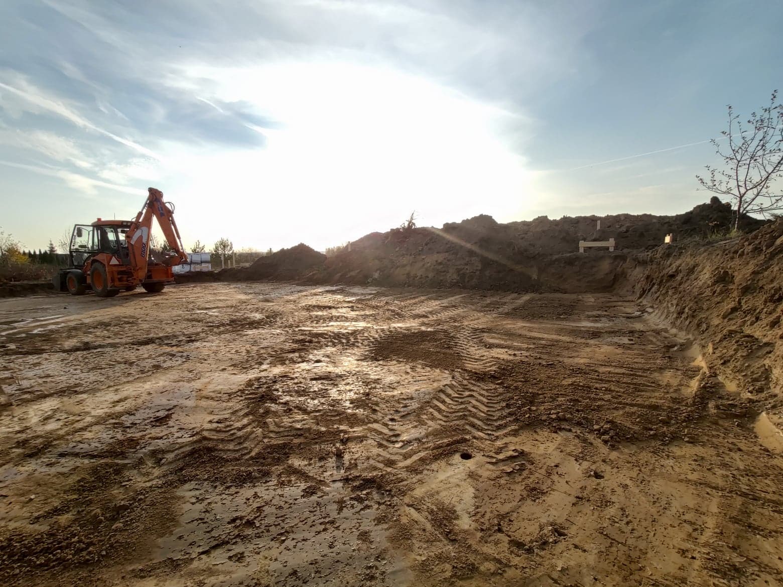 Pomarańczowa koparko-ładowarka na budowie, przygotowany teren z widocznymi śladami opon, wykop z drewnianymi stemplami, w tle hałda ziemi i błękitne niebo z chmurami.