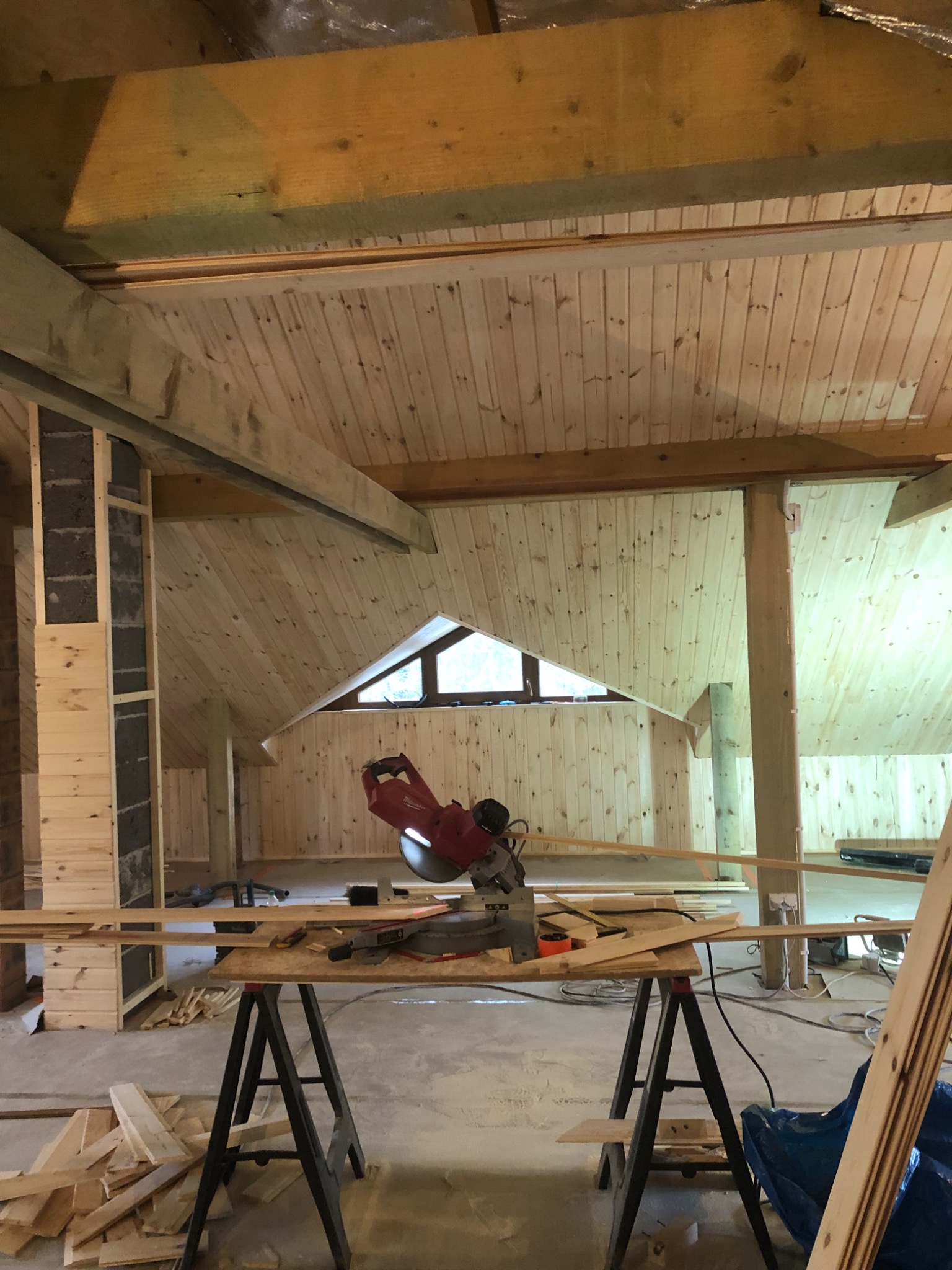 Wnętrze poddasza w trakcie prac wykończeniowych, widoczne drewniane panele ścienne i sufitowe, piła stołowa na statywie, okno w kształcie trójkąta i fragmenty konstrukcji budynku.