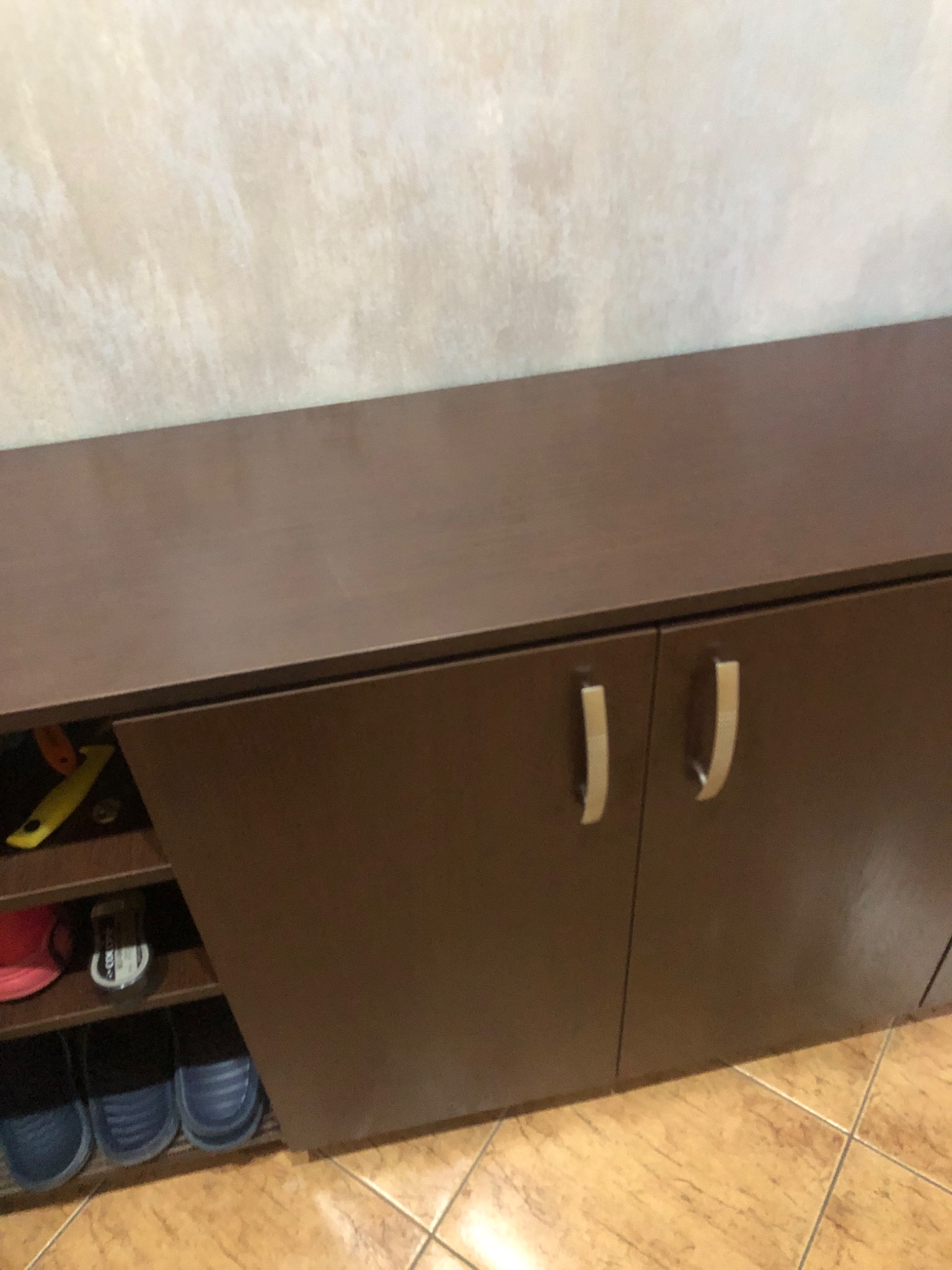 Ciemnobrązowa szafka na buty z metalowymi uchwytami, z otwartymi półkami po lewej stronie, na których widać obuwie i akcesoria, na tle beżowej ściany i podłogi z płytek.