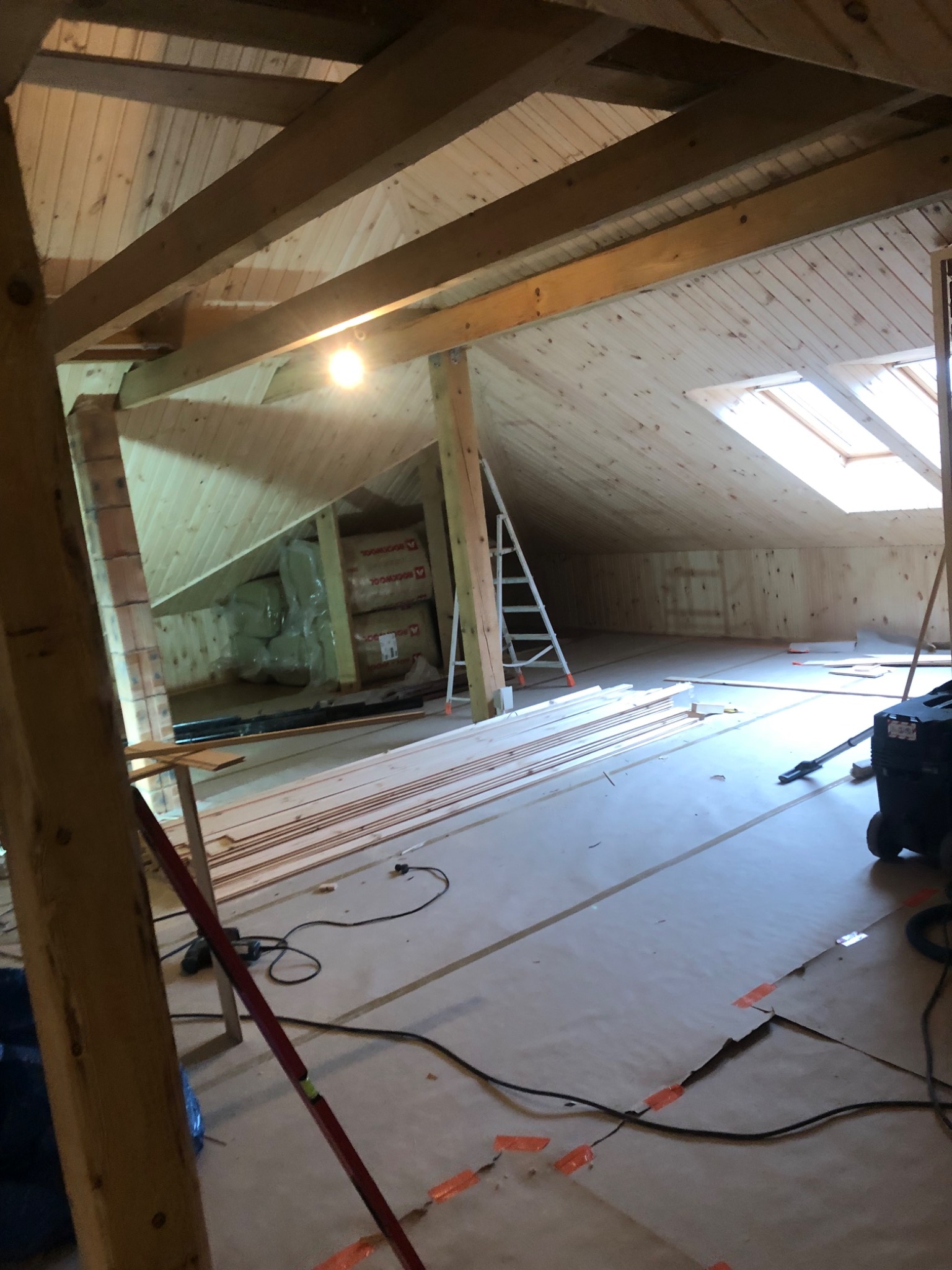 Adaptacja poddasza: drewniana boazeria na ścianach i suficie, widoczne belki konstrukcyjne, okno dachowe, materiały izolacyjne i drabina w tle, podłoga zabezpieczona folią malarską.