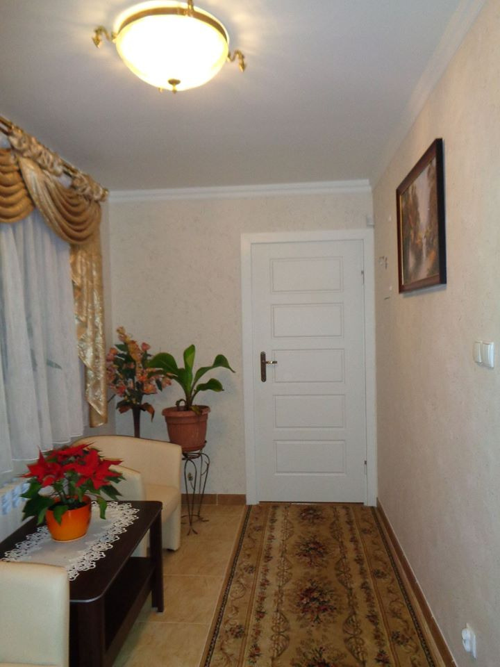 Klasycznie urządzone wnętrze przedpokoju z beżową tapetą o fakturze, białymi drzwiami panelowymi, dywanem w orientalne wzory, kwiatami w doniczkach i lampą sufitową z elementami złota.