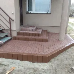Nowo ułożona, czerwono-brązowa kostka brukowa tworząca schody i taras przed wejściem do domu z kolumnami, widoczne metalowe schody z boku.