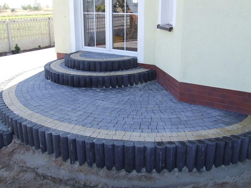 Półokrągłe schody wejściowe z szarej kostki brukowej, obrzeża z ciemnych, okrągłych elementów, jasna linia kostki, elewacja domu w tle, widoczny fragment ogrodu.