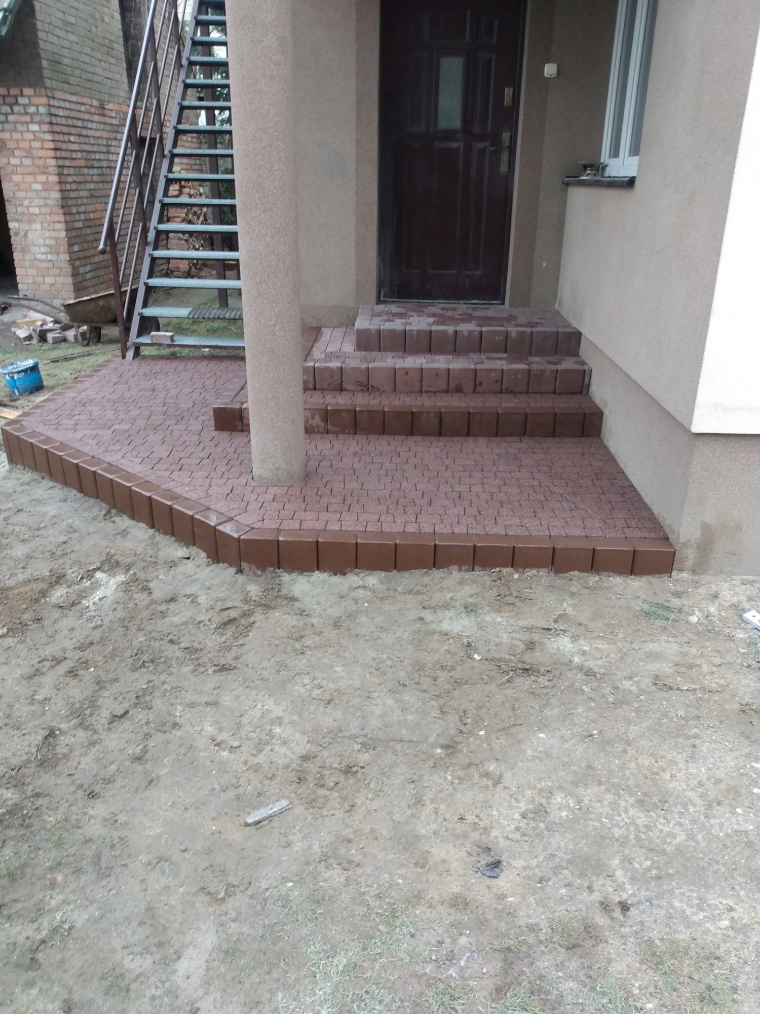 Brązowa kostka brukowa ułożona przed wejściem do domu, schody z kostki prowadzą do ciemnych drzwi, obok kolumna i metalowe schody.