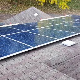 Panele fotowoltaiczne zamontowane na dachu pokrytym brązowym gontem bitumicznym, widoczne elementy mocujące, zabezpieczenia i jesienne otoczenie.