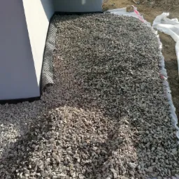 Warstwa drenażowa z kamienia ułożona przy fundamencie budynku, zabezpieczona folią kubełkową i geowł&oacute;kniną, z widocznymi narzędziami w tle.