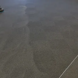 Szary dywan w trakcie czyszczenia z widocznym fragmentem maszyny czyszczącej po lewej stronie, na dywanie widoczne ślady czyszczenia w postaci jaśniejszych plam, tło stanowi puste pomieszczenie...