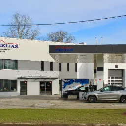 Budynek stacji kontroli pojazdów ZELIAS z dwoma samochodami osobowymi zaparkowanymi przy dystrybutorze AdBlue, widoczne trzy bramy garażowe z numerami.