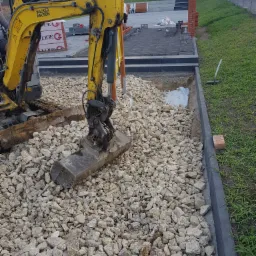 Żółta mini koparka Wacker Neuson na placu budowy, wysypująca biały żwir na geowłókninę w przygotowanym wykopie pod nawierzchnię, obok widoczny krawężnik i trawnik.