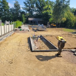 FUH MARIO-BUD - Żółta ubijarka wibracyjna stoi na utwardzanej żwirowej powierzchni, w tle wykop z obrzeżami, teren budowy z maszynami i zielenią.