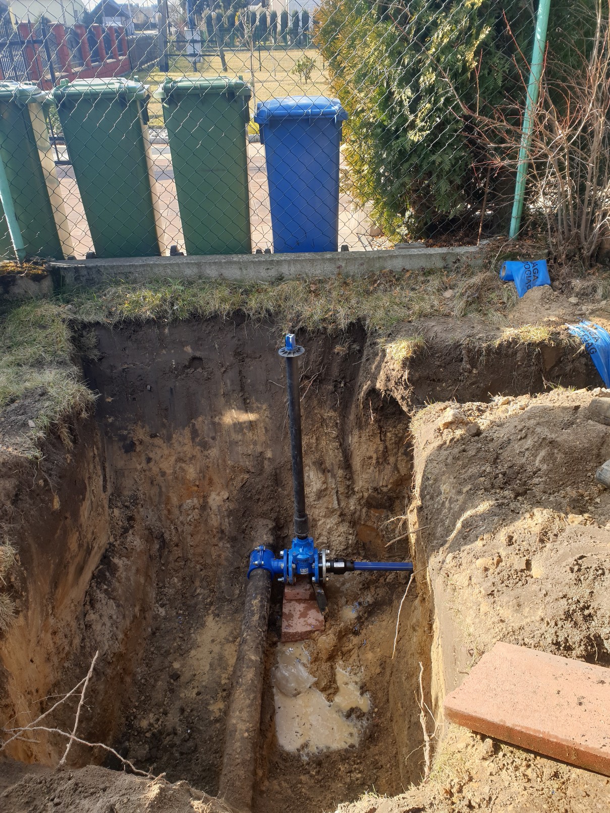 Głęboki wykop w ziemi, w którym widoczne są niebieskie zawory i rury instalacji wodnej, wsparte na cegłach, z trzema zielonymi i jednym niebieskim pojemnikiem na śmieci w tle.