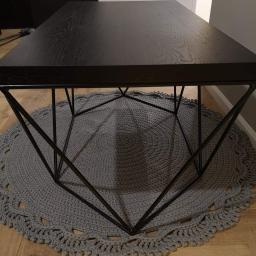 Avanti Design - Niski stolik kawowy z czarnym, drewnianym blatem i geometryczną, metalową podstawą, ustawiony na okrągłym, szarym dywaniku z dzianiny.