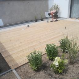 Avanti Design - Mężczyzna w czapce maluje drewniany taras z jasnych desek na zewnątrz domu z elementami ogrodu i roślinami.