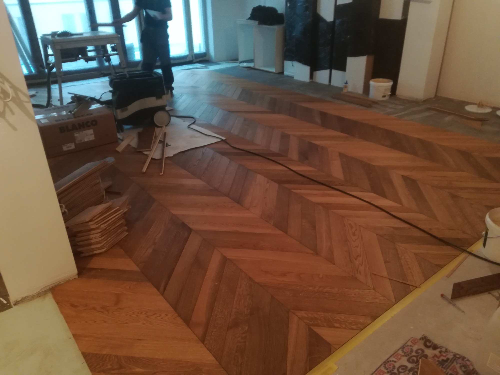 Układanie parkietu w jodełkę z widocznymi narzędziami i materiałami budowlanymi w trakcie pracy.