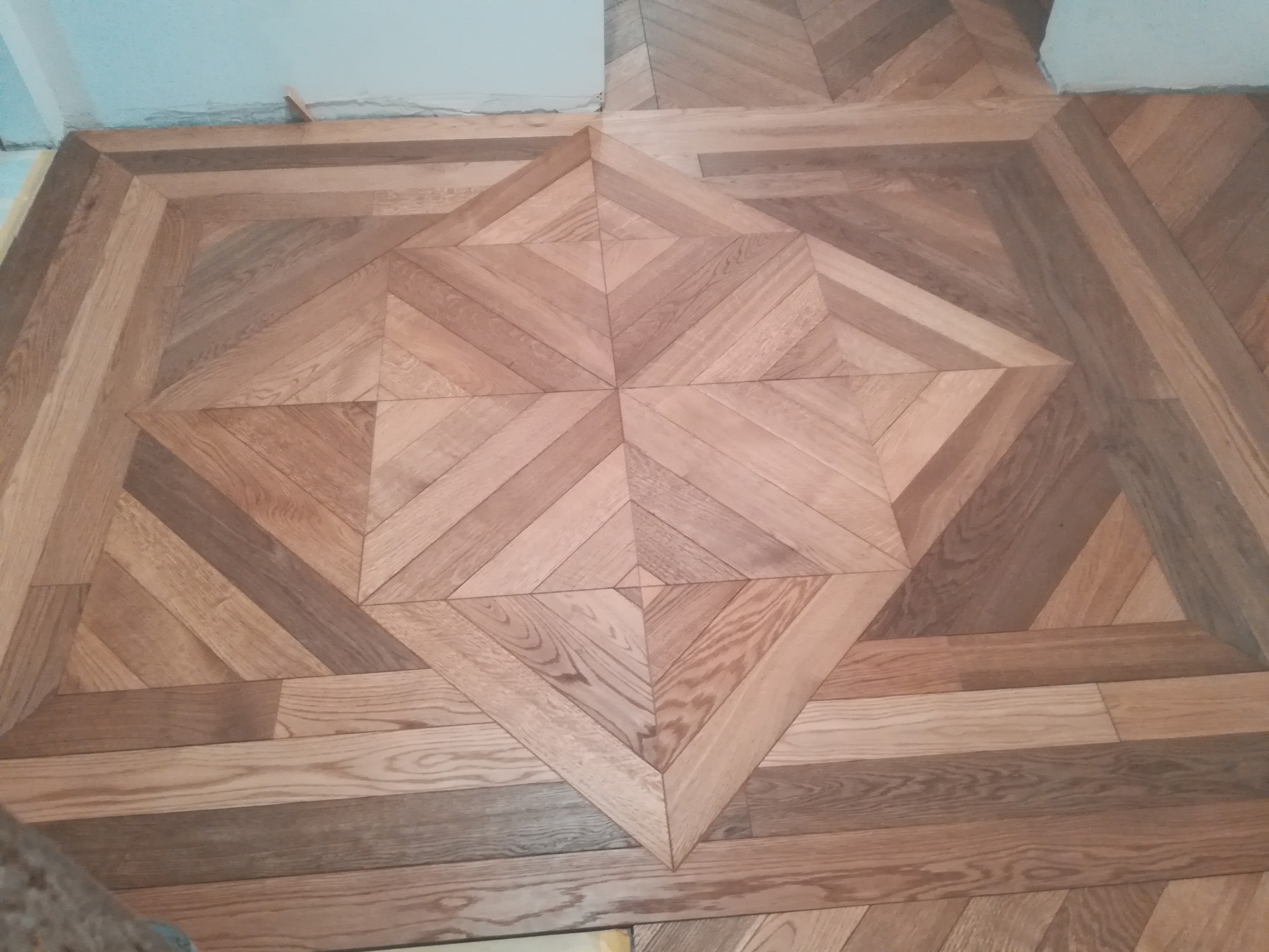 Fragment drewnianej podłogi z parkietem ułożonym we wzór gwiazdy, widoczne niedoskonałości wykończenia przy ścianie.