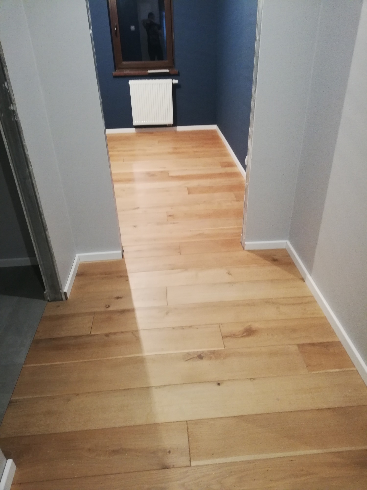 Jasna drewniana podłoga ułożona w korytarzu z szarymi ścianami i widokiem na pokój z ciemnoniebieskimi ścianami, białym grzejnikiem i oknem.