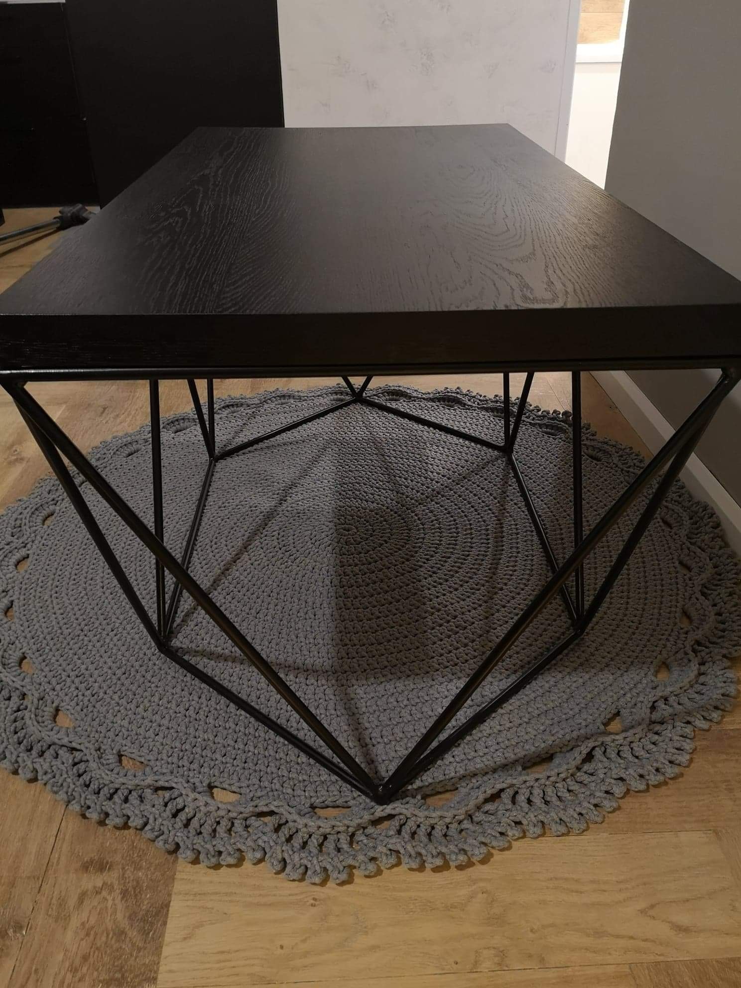 Niski stolik kawowy z czarnym, drewnianym blatem i geometryczną, metalową podstawą, ustawiony na okrągłym, szarym dywaniku z dzianiny.
