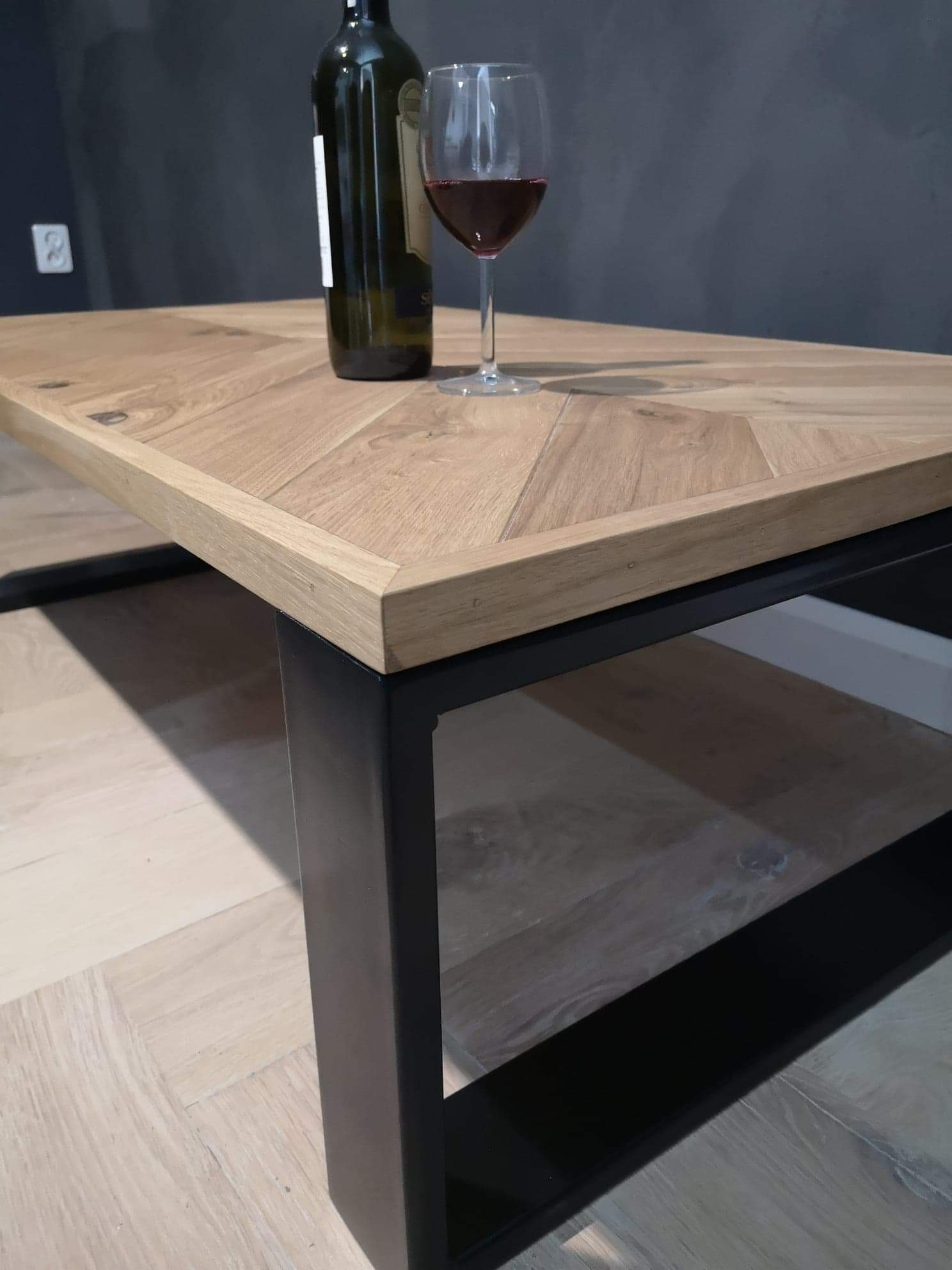 Stolik kawowy z drewnianym blatem ułożonym we wzór jodełki, na metalowych, czarnych nogach, z butelką wina i kieliszkiem na blacie.