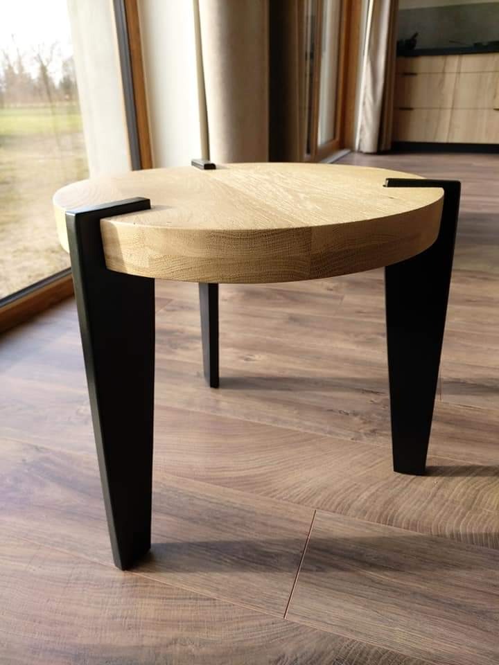 Okrągły stolik kawowy z jasnego drewna z czarnymi, stożkowymi nogami na drewnianej podłodze w jasnym wnętrzu.