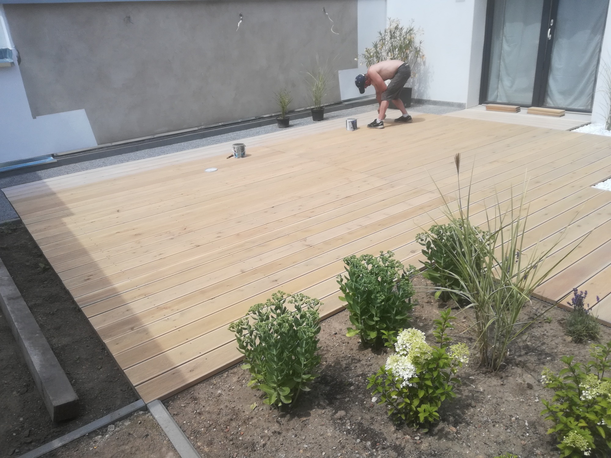 Mężczyzna w czapce maluje drewniany taras z jasnych desek na zewnątrz domu z elementami ogrodu i roślinami.