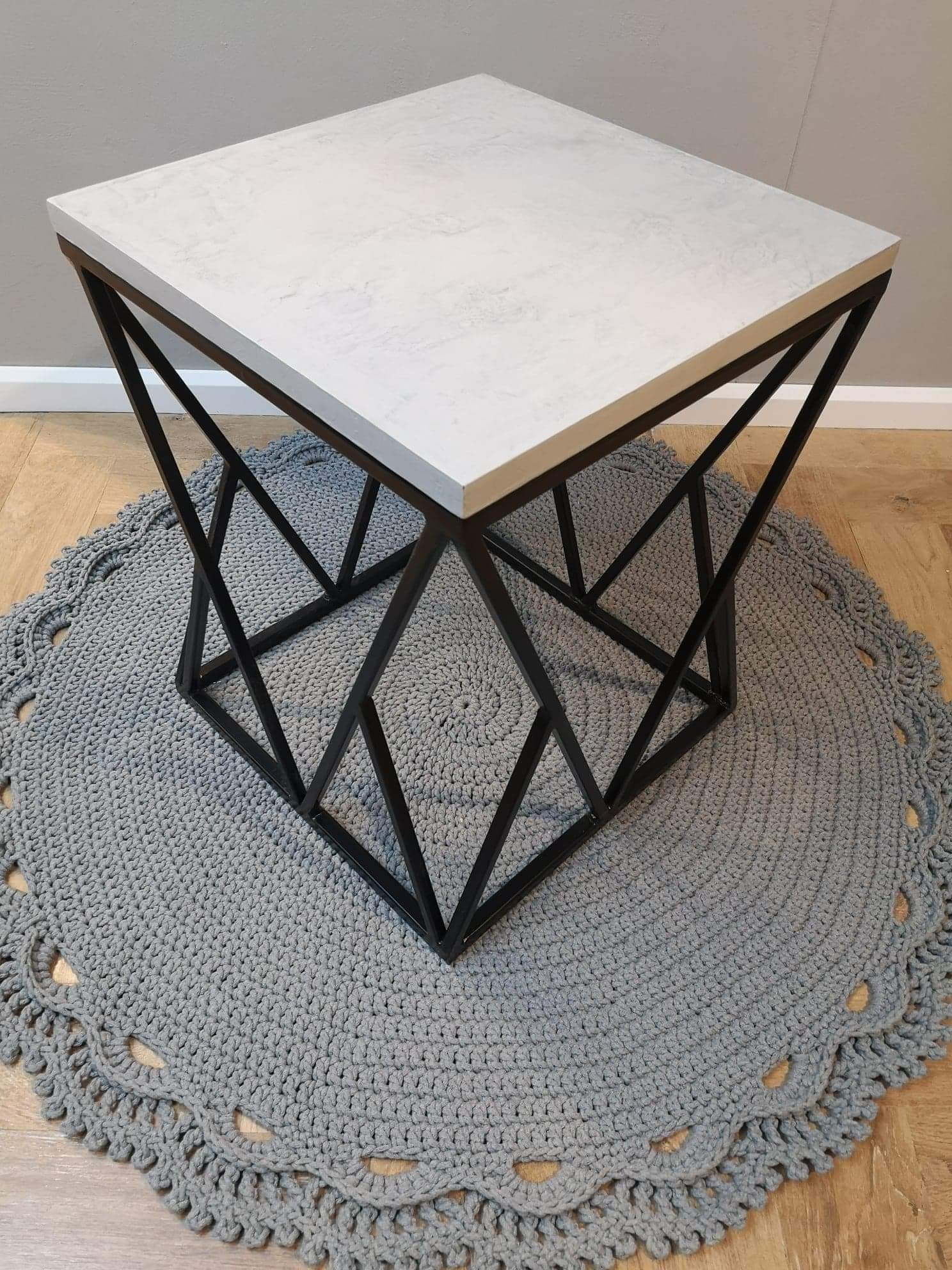 Stolik kawowy z blatem imitującym beton, osadzony na geometrycznej, metalowej podstawie, leżący na okrągłym, szarym dywanie ze sznurka.