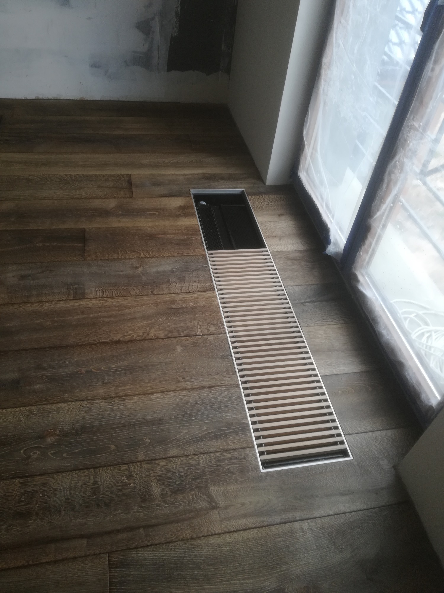 Ciemna, drewniana podłoga z wbudowanym, liniowym odpływem podłogowym ze stali nierdzewnej, widoczne okno zabezpieczone folią malarską.