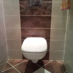 Wisząca toaleta w łazience z brązowo-beżowymi płytkami ułożonymi w geometryczny wzór, widoczny panel spłukujący i fragment ręcznika.