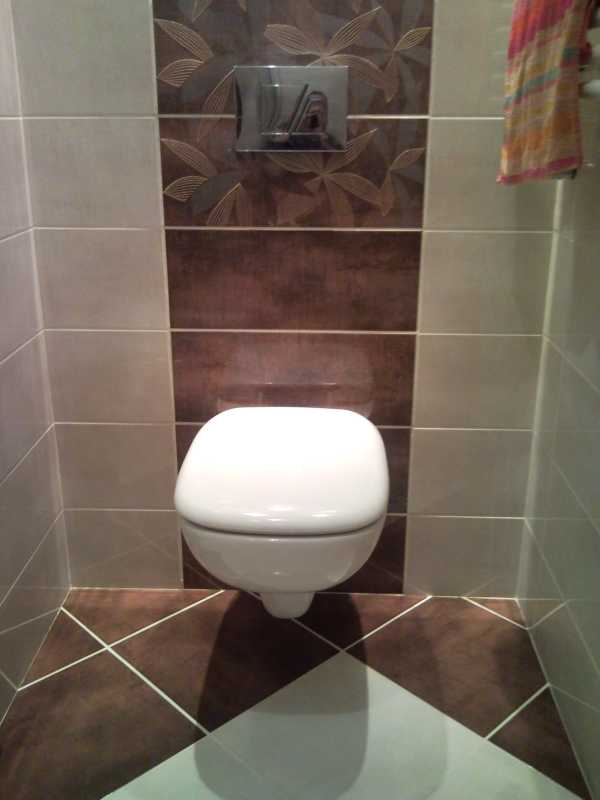 Wisząca toaleta w łazience z brązowo-beżowymi płytkami ułożonymi w geometryczny wzór, widoczny panel spłukujący i fragment ręcznika.