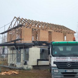 DREW-DOM Andrzej Antecki - Wznoszenie konstrukcji domu szkieletowego z widocznym drewnianym szkieletem, płytami OSB, rusztowaniami, dźwigiem i zaparkowaną ciężarówką z napisem 'Hurtownia Budowlana' na tle pochmurnego nieba.
